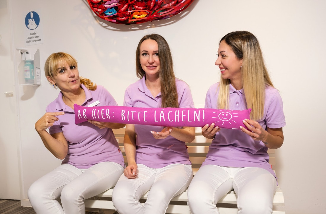 Lächelnde Zahnarzthelferinnen halten ein Schild mit der Aufschrift „Ab hier: Bitte lächeln!“ in der Praxis.